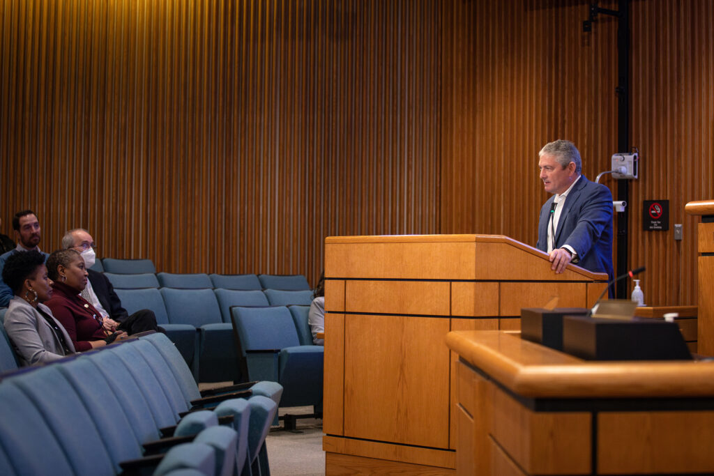 View from the right side of the Board Room of the County Manager at the podium and Yvette George and Monica Callahan-Smith watching him from the second row