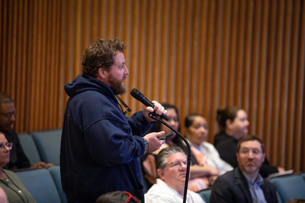 Right profile view of a DPU employee speaking into a microphone and holding his phone among the seats of the audience.
