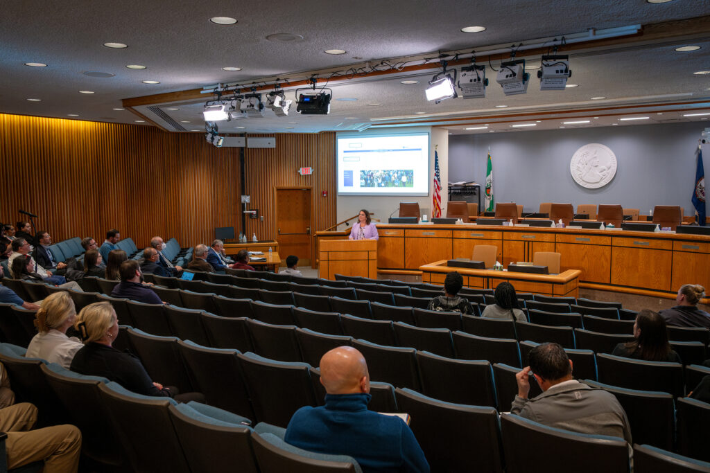 Wide room view of the Board Room with Sheila Minor at the podium, in front of a screen. Employees are scattered throughout the audience seats